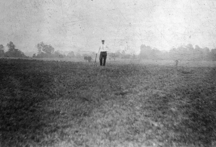 Black and white photograph of a solitary figure in light-colored clothing standing in a vast, flat agricultural field with scattered trees along the horizon.