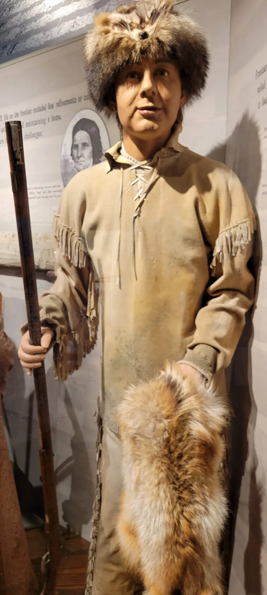 Museum display mannequin wearing traditional frontier clothing including a tan leather tunic with fringe detailing, fur-lined boots, and a fur hat. The figure holds a long rifle and stands against a white wall with historical text and a portrait photograph visible in the background.