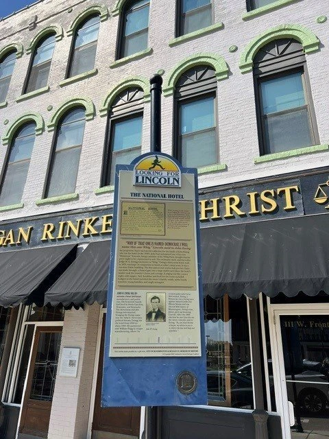 Historic brick building facade with arched windows framed in green, featuring a commemorative plaque for The National Hotel mounted on the storefront. Ground-level storefronts display partial signage including 