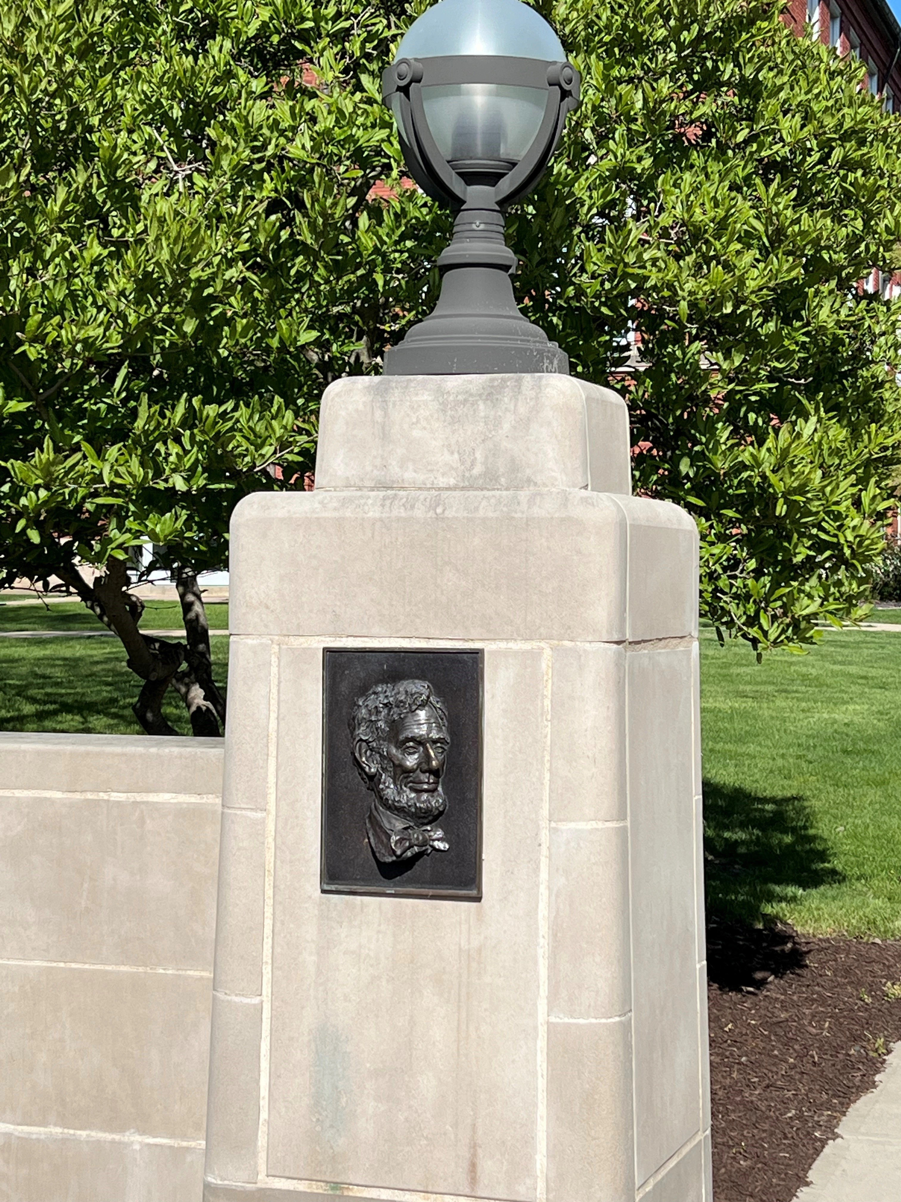 A stone pedestal monument topped with a black lamp post, featuring a dark bronze relief portrait plaque of a bearded man wearing a bow tie. The monument is set against a backdrop of green hedges and manicured lawn.