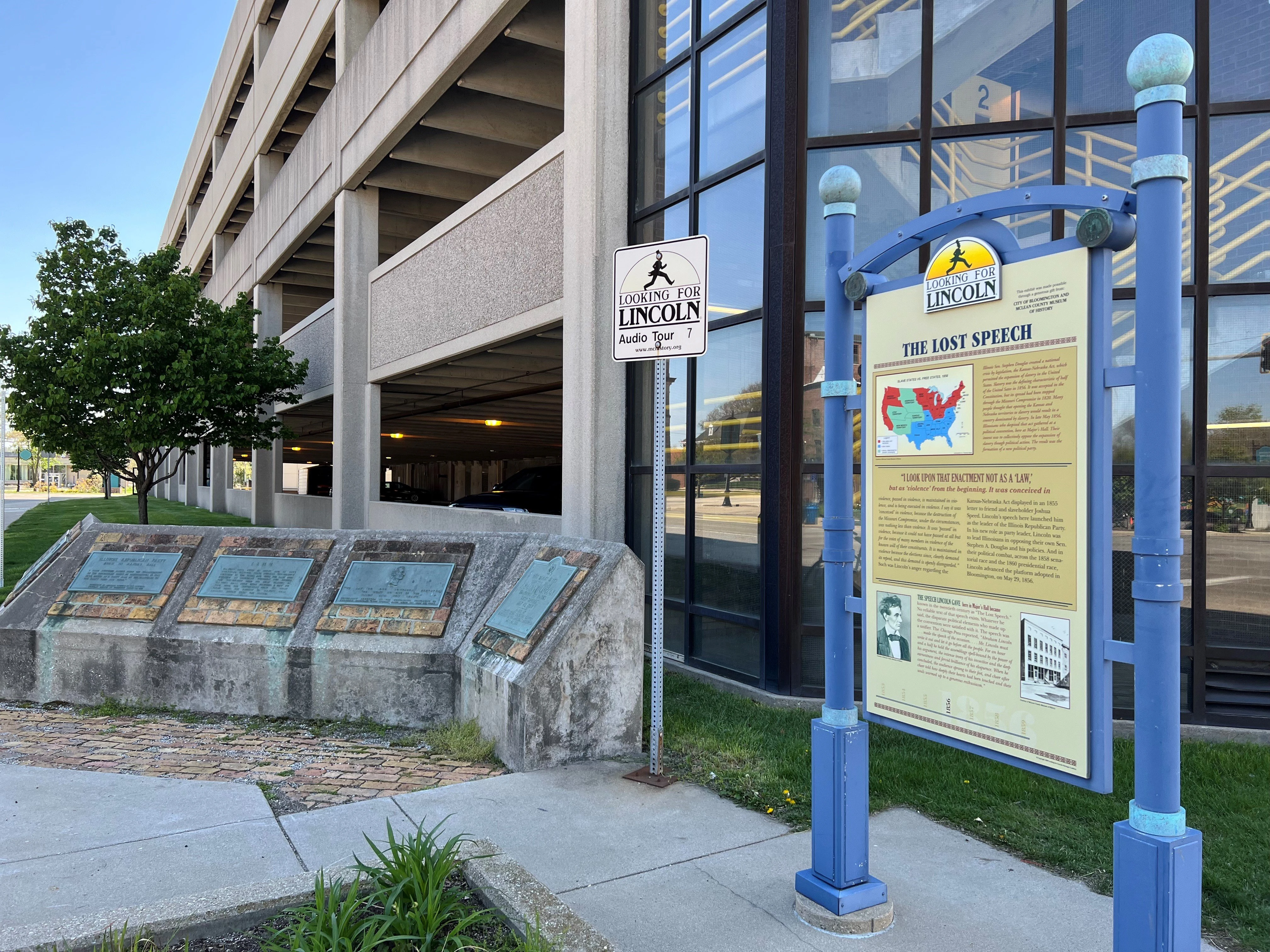 Outdoor historical marker display mounted on blue poles next to a concrete building. A yellow and blue informational sign reads 