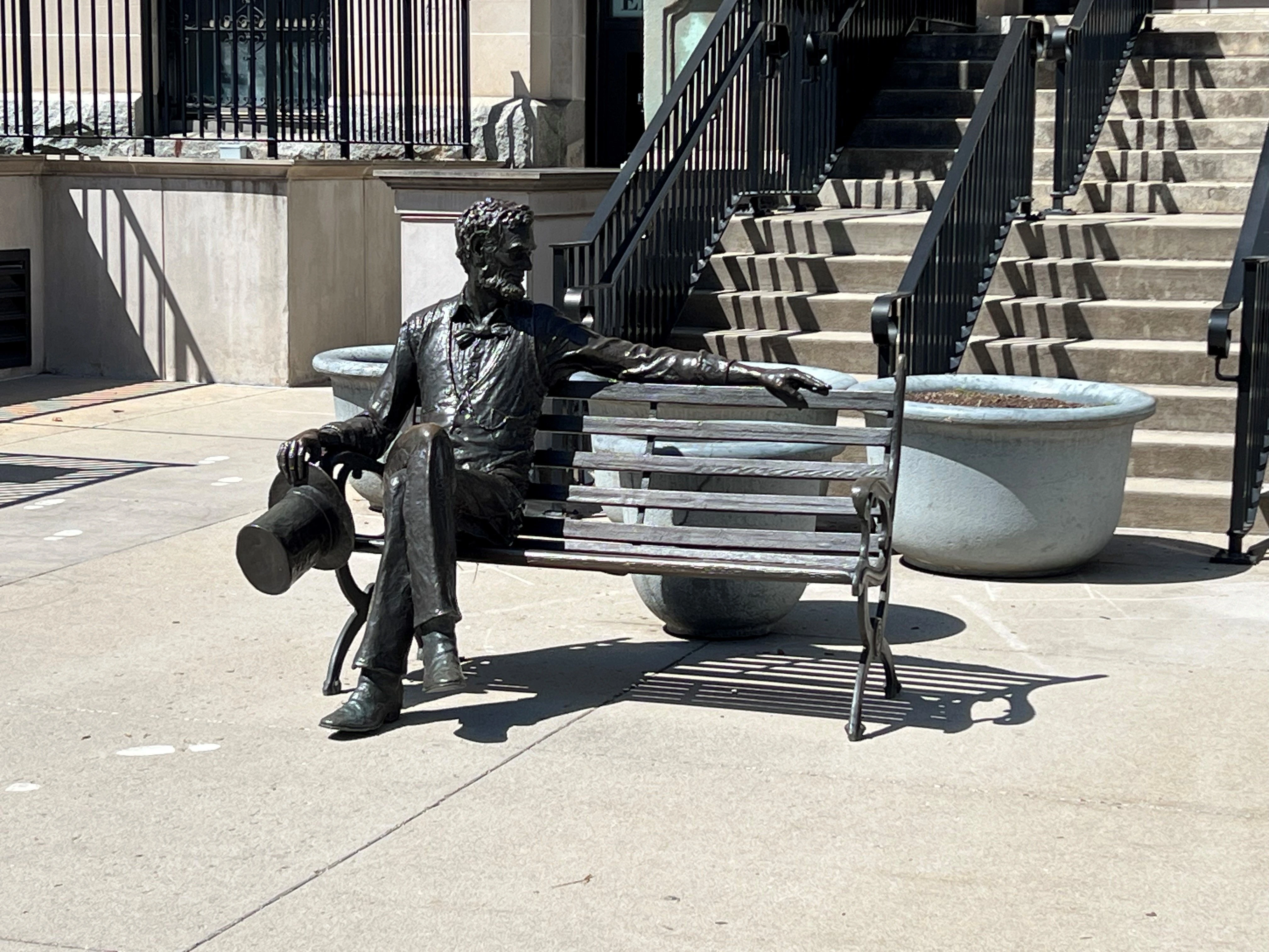 Bronze statue of a bearded man in a suit sitting on a bench in a public plaza, holding a hat in one hand with his arm extended, surrounded by stone planters and wide stairs with black railings in the background.