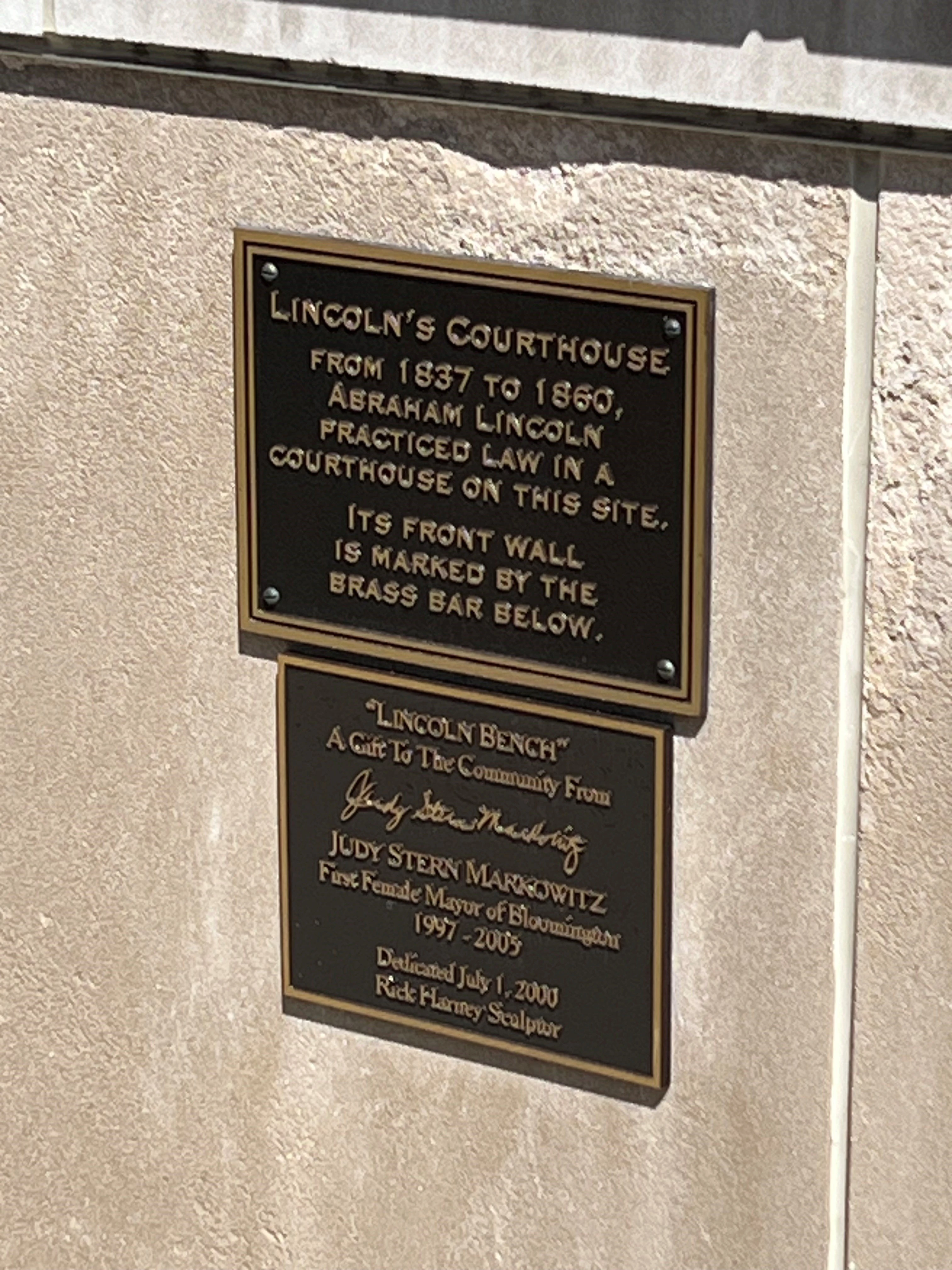 Two brass plaques mounted on a textured stone wall. The upper plaque commemorates Lincoln's Courthouse from 1837 to 1860, where Abraham Lincoln practiced law, with its front wall marked by a brass bar below. The lower plaque dedicates a 