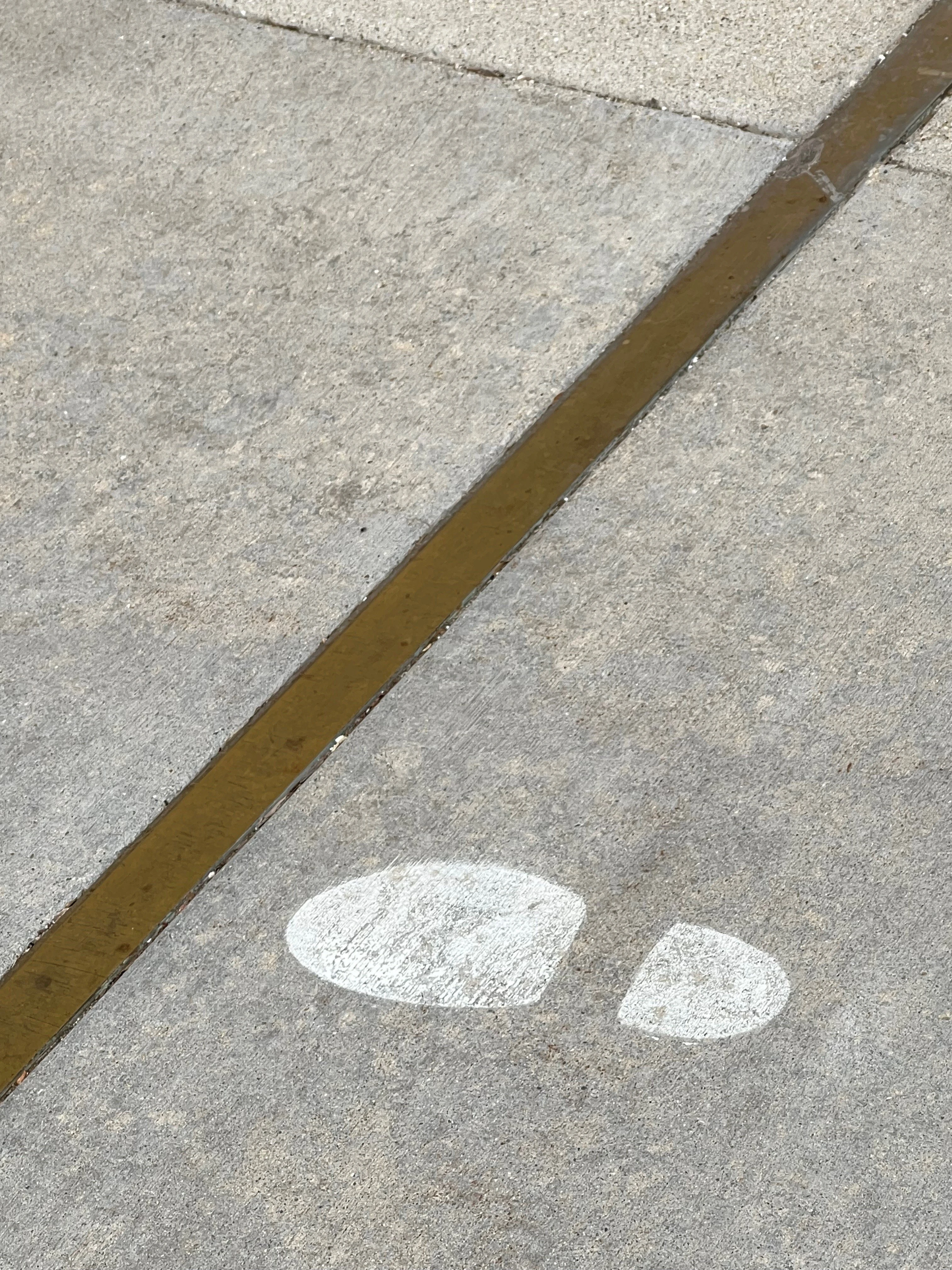 Concrete pavement with a metal expansion joint strip running diagonally across it, and two white painted footprints marked on the surface below the joint.