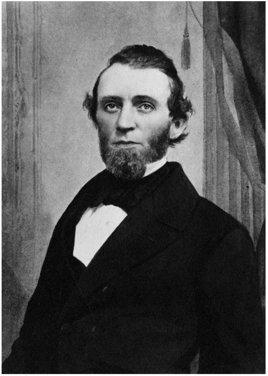 Black and white portrait photograph of a bearded man in formal 19th century attire, wearing a dark jacket with white shirt front, facing slightly toward the camera against a draped curtain background.