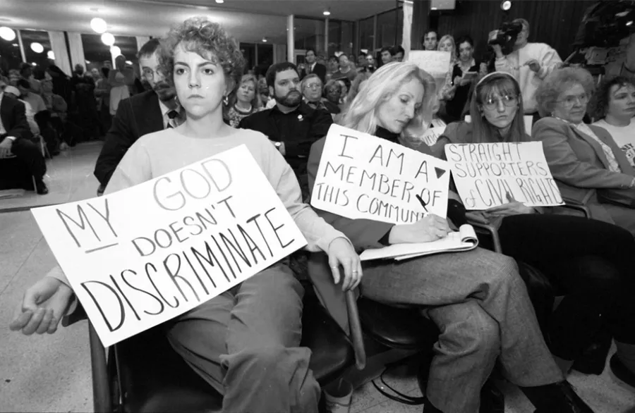 Women sit with poster board signs saying My God Doesn't Discriminate, and I Am a Member of This Community.