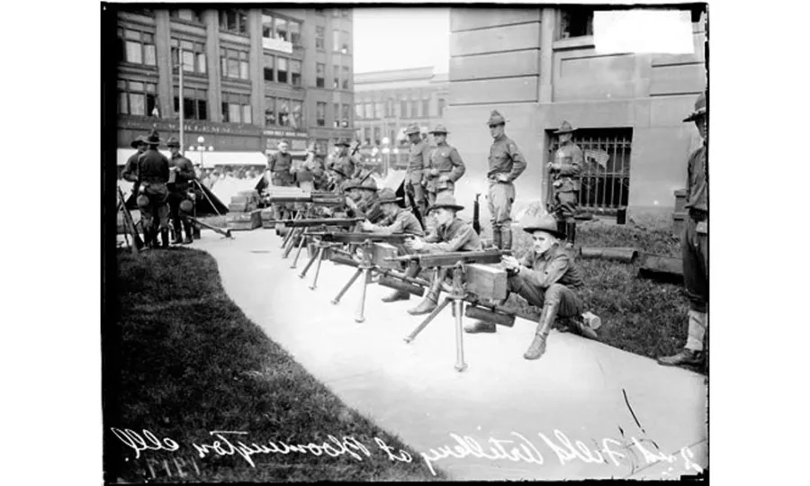 A group of soldiers in large hats sit behind a line of machine guns along a sidewalk. Another group stands behind them with hands on hips.