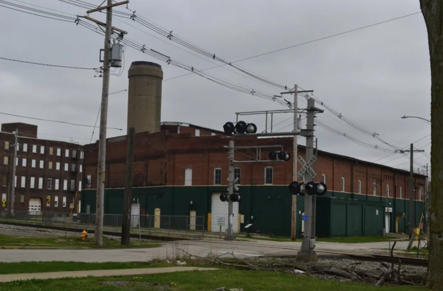 Two story brick building with smokestack and large garage doors adjacent to a rail line and power lines.