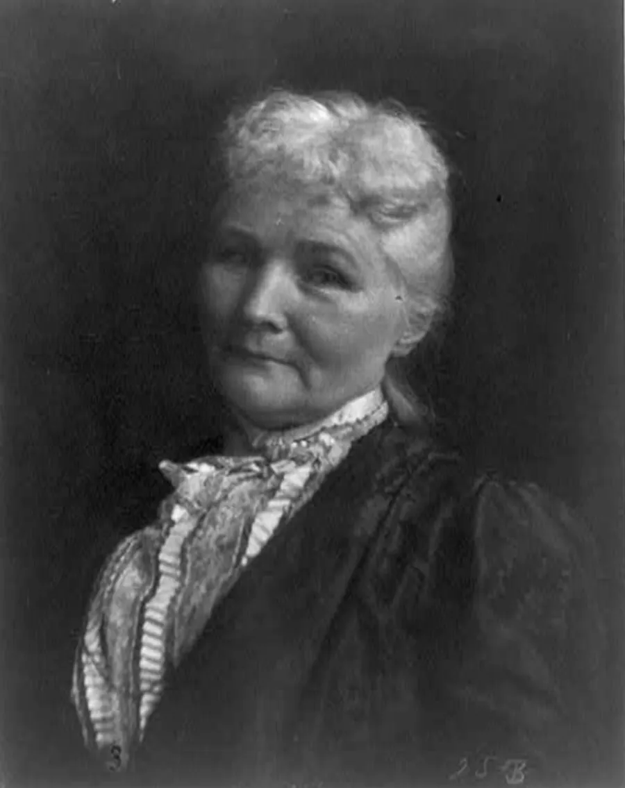 Black and white portrait of a middle-aged woman looking at the camera. She has a sharp nose, white hair in a bun, and is wearing a dark blouse with ruffled collar.