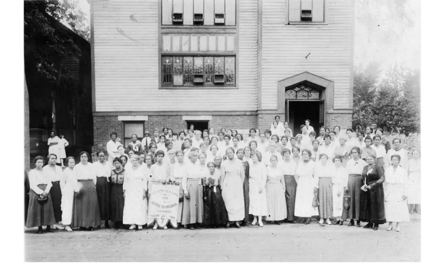 Black and white photo of more than 100 women in dresses posing in front of a building with white siding.