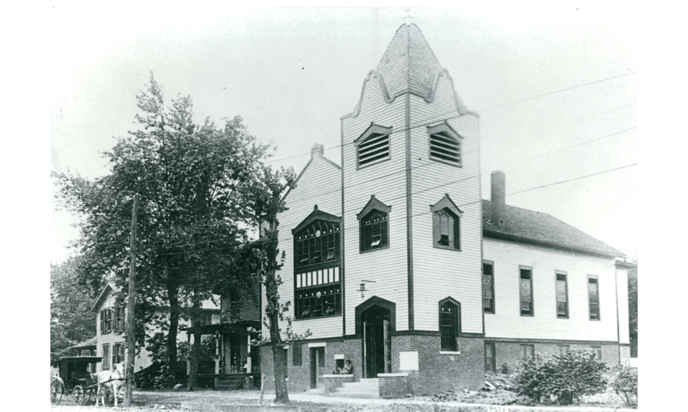 Brick and wood siding church with a three-story steeple and one story hall.