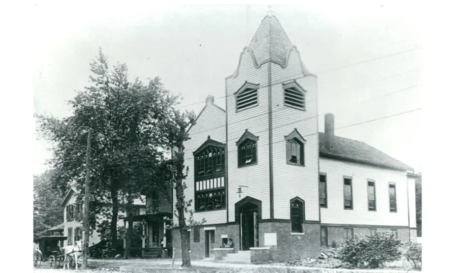 Brick and wood siding church with a three-story steeple and one story hall.