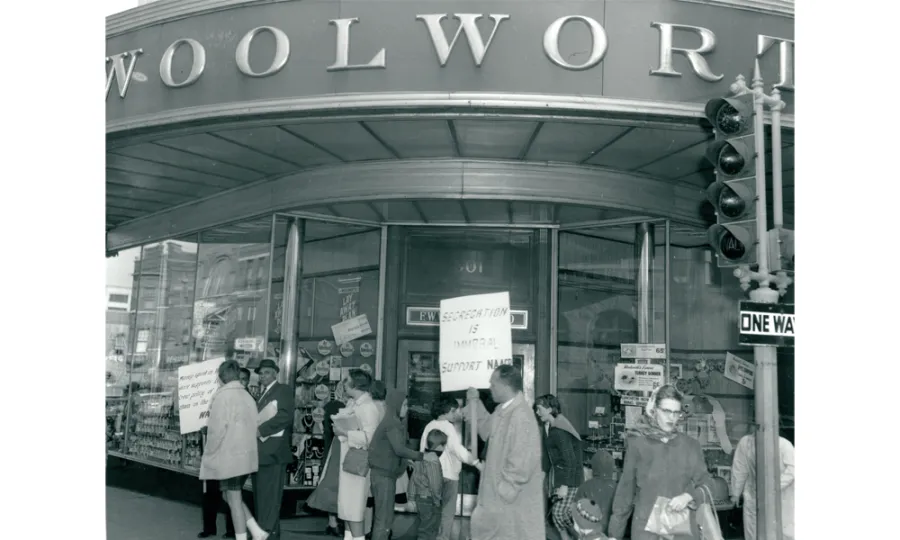 Black and white photograph of a protest or picket line at Woolworth storefront, with multiple people holding signs, circa 1960s civil rights era.
