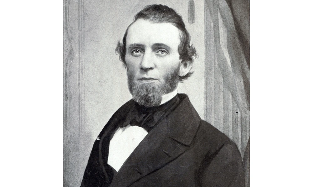 Black and white portrait photograph of a bearded man in formal 19th-century attire, wearing a dark suit jacket and white dress shirt with bow tie, posed against a draped curtain backdrop.