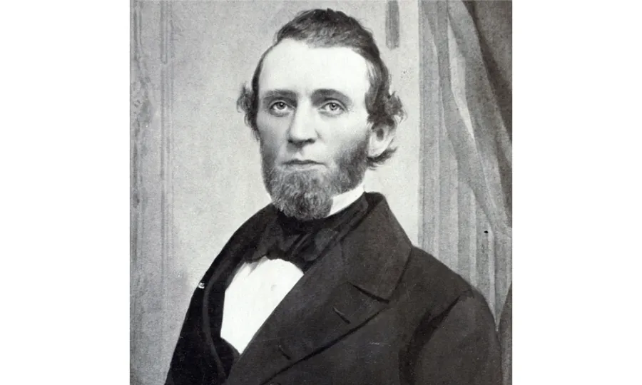 Black and white portrait photograph of a bearded man in formal 19th-century attire, wearing a dark suit jacket and white dress shirt with bow tie, posed against a draped curtain backdrop.