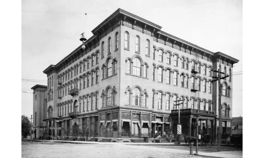 Black and white photograph of a large four-story commercial building with ornate architectural details, featuring numerous arched windows arranged in regular rows across its facade. The ground floor contains storefronts with display windows. A decorative cornice and balustrade crown the roofline. Period utility poles and a dirt street are visible in the foreground.