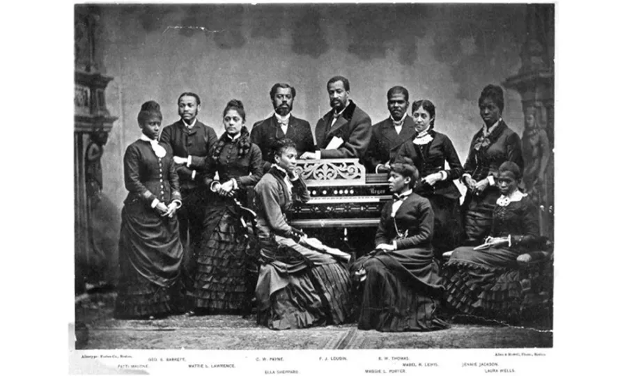 Black and white photograph of a Victorian-era group portrait featuring twelve people arranged around an ornate harmonium or pump organ. Eight men stand in the back row wearing dark suits, while four women are seated in the front, dressed in dark Victorian attire with full skirts. The photograph appears to be a formal studio portrait with names listed at the bottom identifying the subjects.