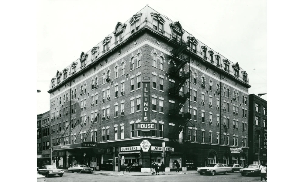 Black and white photograph of a distinctive corner building with mansard roof and dormer windows, featuring ground-floor storefronts including a jewelry store and house of ill repute sign, with vintage automobiles parked on the street, circa mid-20th century.