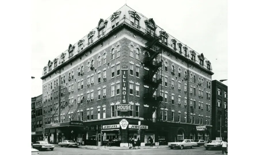 Black and white photograph of a distinctive corner building with mansard roof and dormer windows, featuring ground-floor storefronts including a jewelry store and house of ill repute sign, with vintage automobiles parked on the street, circa mid-20th century.