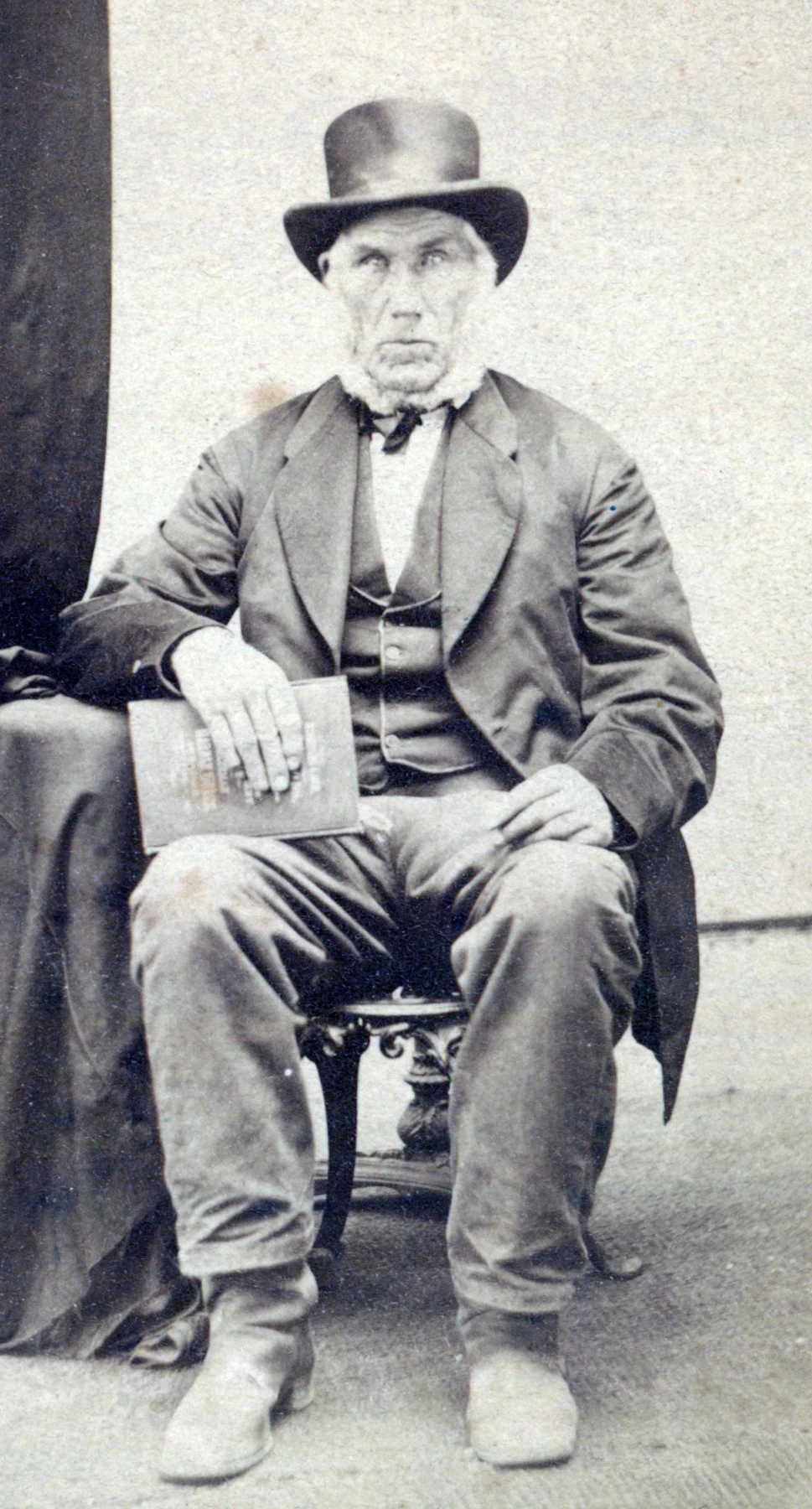 A white man wearing a three piece suit and a tophat. he has a thick white beard that goes around the outside edges of his face and below the chin, but the rest of his face is clean shaven.