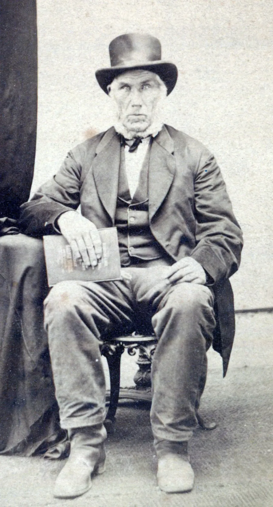 A white man wearing a three piece suit and a tophat. he has a thick white beard that goes around the outside edges of his face and below the chin, but the rest of his face is clean shaven.