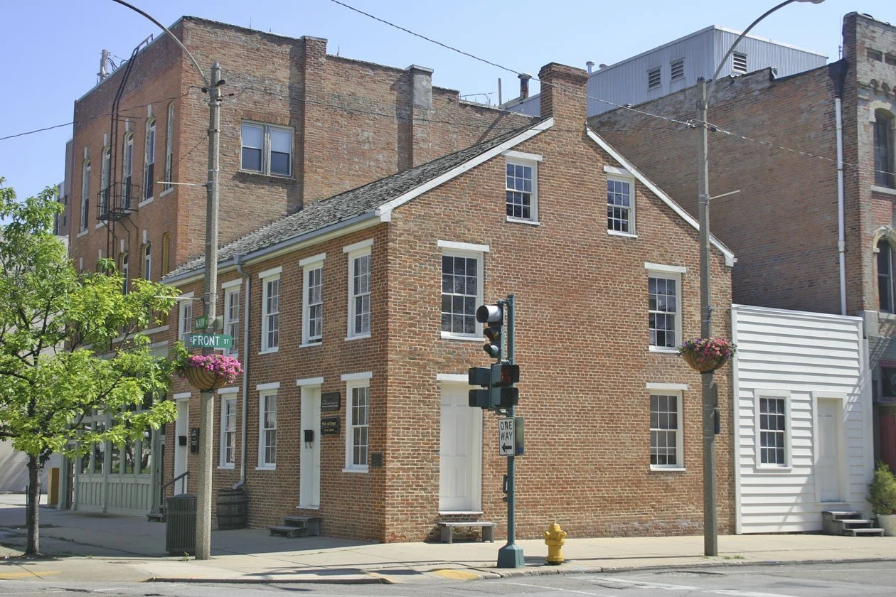 Historic brick buildings at a street corner intersection, featuring a prominent two-story red brick structure with white-trimmed windows and a gabled roof, adjacent to taller commercial buildings, with a traffic light and flowering baskets visible along the street.