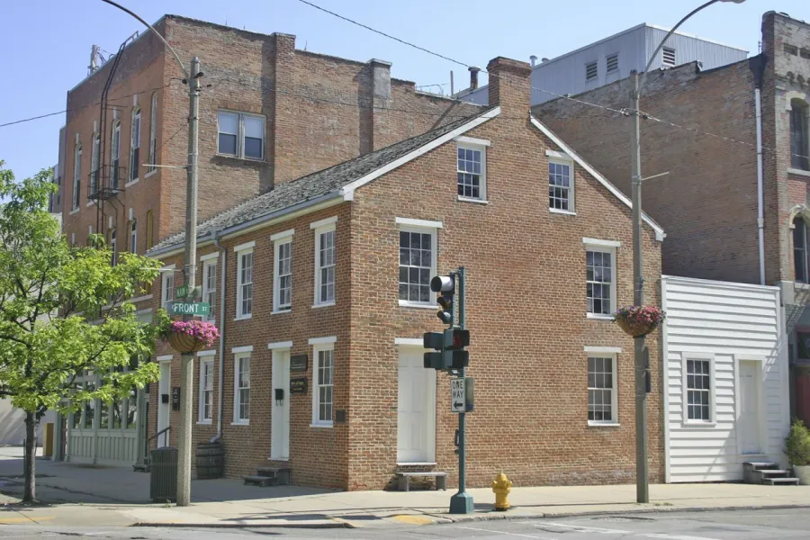 Historic brick buildings at a street corner intersection, featuring a prominent two-story red brick structure with white-trimmed windows and a gabled roof, adjacent to taller commercial buildings, with a traffic light and flowering baskets visible along the street.