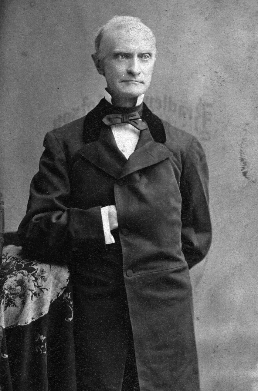 a white man wearing a suit and long coat. His coat is buttoned and his right hand is resting in between the buttons over his chest. he is looking off to the side. he has a straight face and short light colored hair.