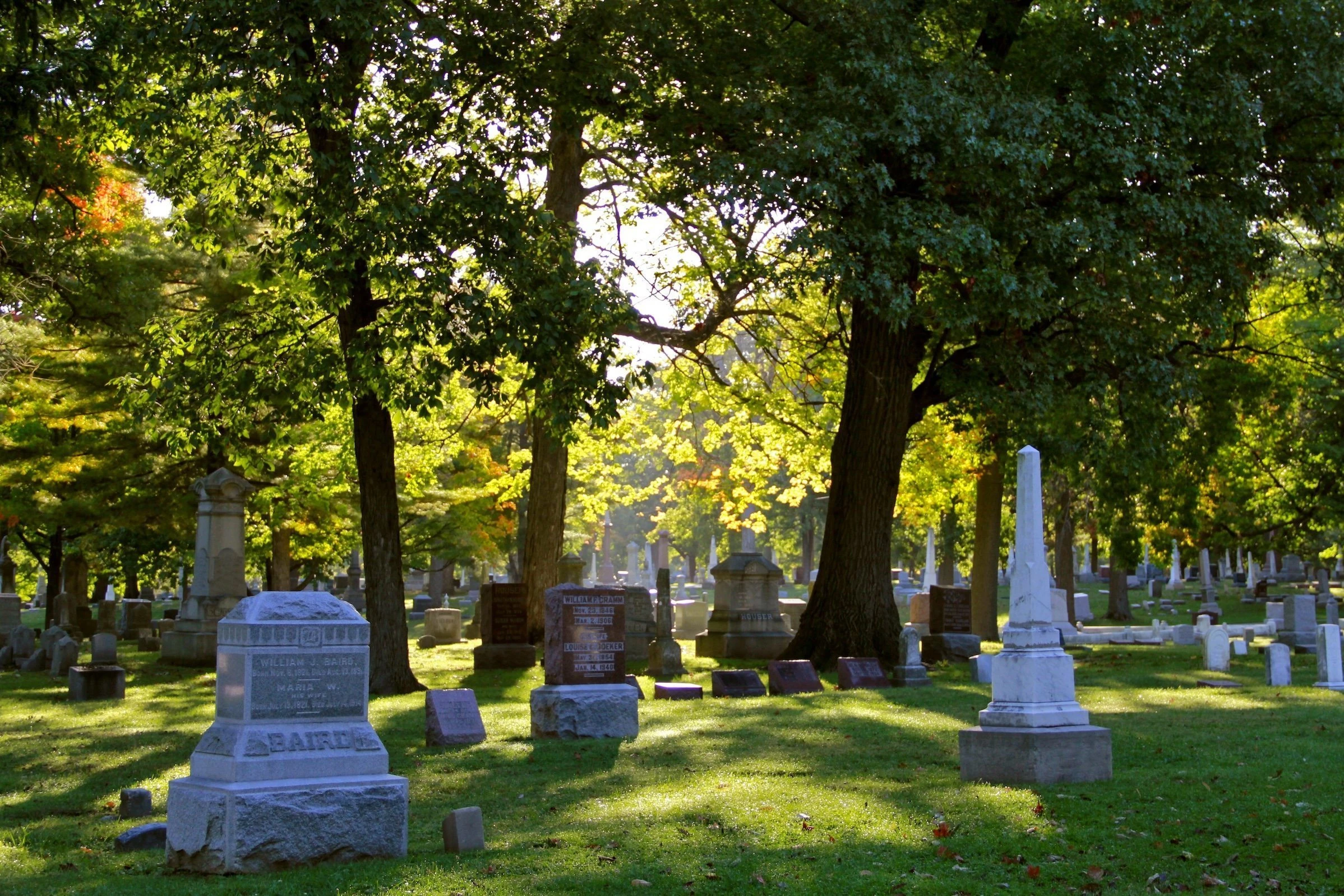 A historic cemetery with numerous stone monuments and headstones scattered across a grassy lawn beneath tall, leafy trees. Dappled sunlight filters through the green foliage, creating patterns of light and shadow across the grounds. Various grave markers of different sizes and styles are visible throughout the peaceful, tree-lined burial ground.