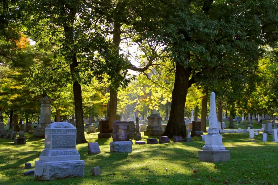 A historic cemetery with numerous stone monuments and headstones scattered across a grassy lawn beneath tall, leafy trees. Dappled sunlight filters through the green foliage, creating patterns of light and shadow across the grounds. Various grave markers of different sizes and styles are visible throughout the peaceful, tree-lined burial ground.
