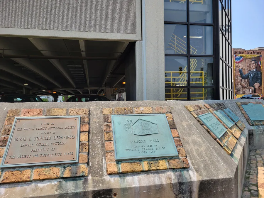 Historical plaques mounted on a brick ledge beneath a concrete overpass. The left plaque honors Wayne C. Townley (1894-1966), a citizen historian. The center turquoise plaque marks Majors Hall, erected in 1829 by William Bradie Major, dated 1920. Additional plaques visible on the right. Modern building with black-framed windows and a mural depicting Lincoln's Lost Speech are visible in the background.