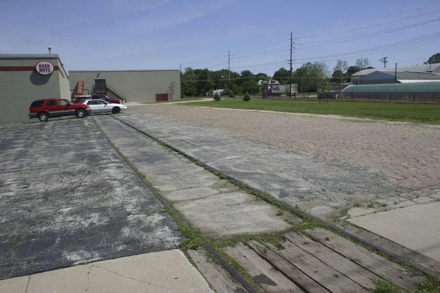 Abandoned parking lot with cracked asphalt and weeds, adjacent to a Beer Nuts building with two parked cars, surrounded by green grass, utility poles, and commercial structures under a clear blue sky.