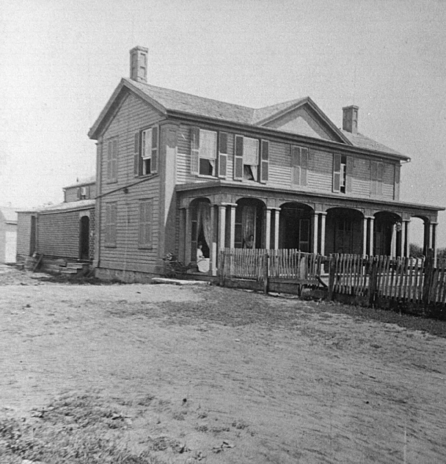 Black and white photograph of a two-story wooden house with horizontal siding, featuring a front porch with columns and white railings, two chimneys, and multiple windows. The house sits on bare ground with a small outbuilding visible to the left.