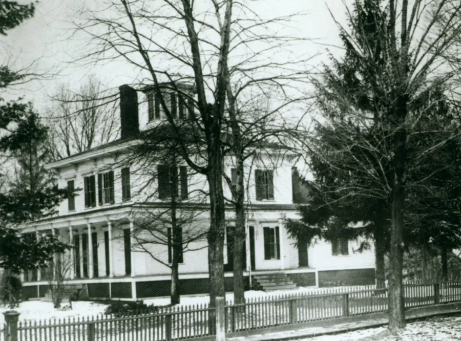 White two-story house with black shutters surrounded by bare trees and white picket fence during winter, with brick chimney visible on roofline.