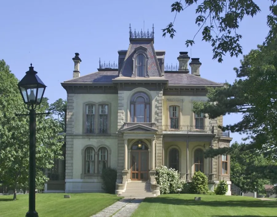 A grand Victorian mansion with pale green stone facade, mansard roof with dormer windows, ornate chimneys, and a columned front entrance. The house is surrounded by manicured lawns and mature trees, with a black vintage street lamp in the foreground.