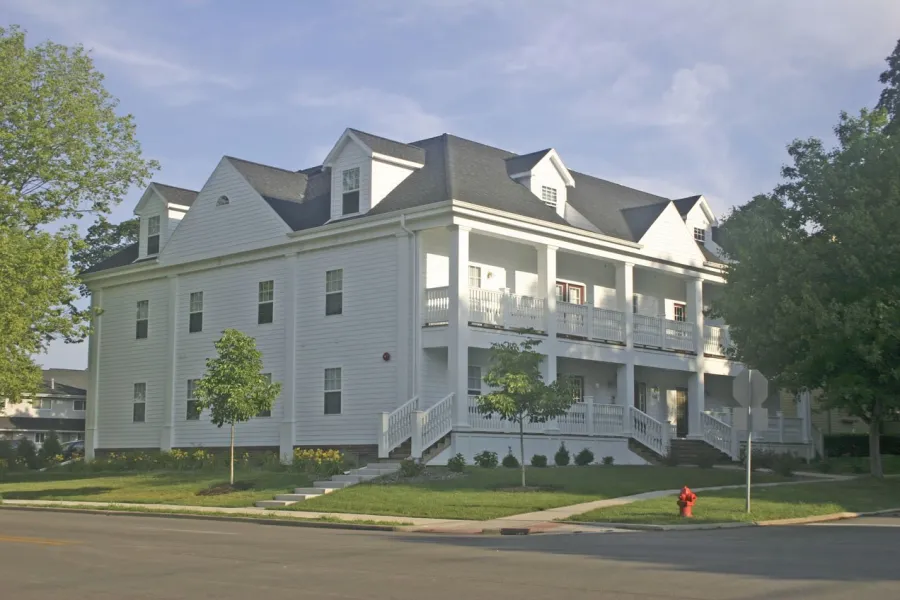 Large white colonial-style house with multiple gables and dormers, featuring wraparound porches on two levels with white railings. The three-story structure sits on a corner lot with mature trees, manicured lawn, and a red fire hydrant visible in the foreground.